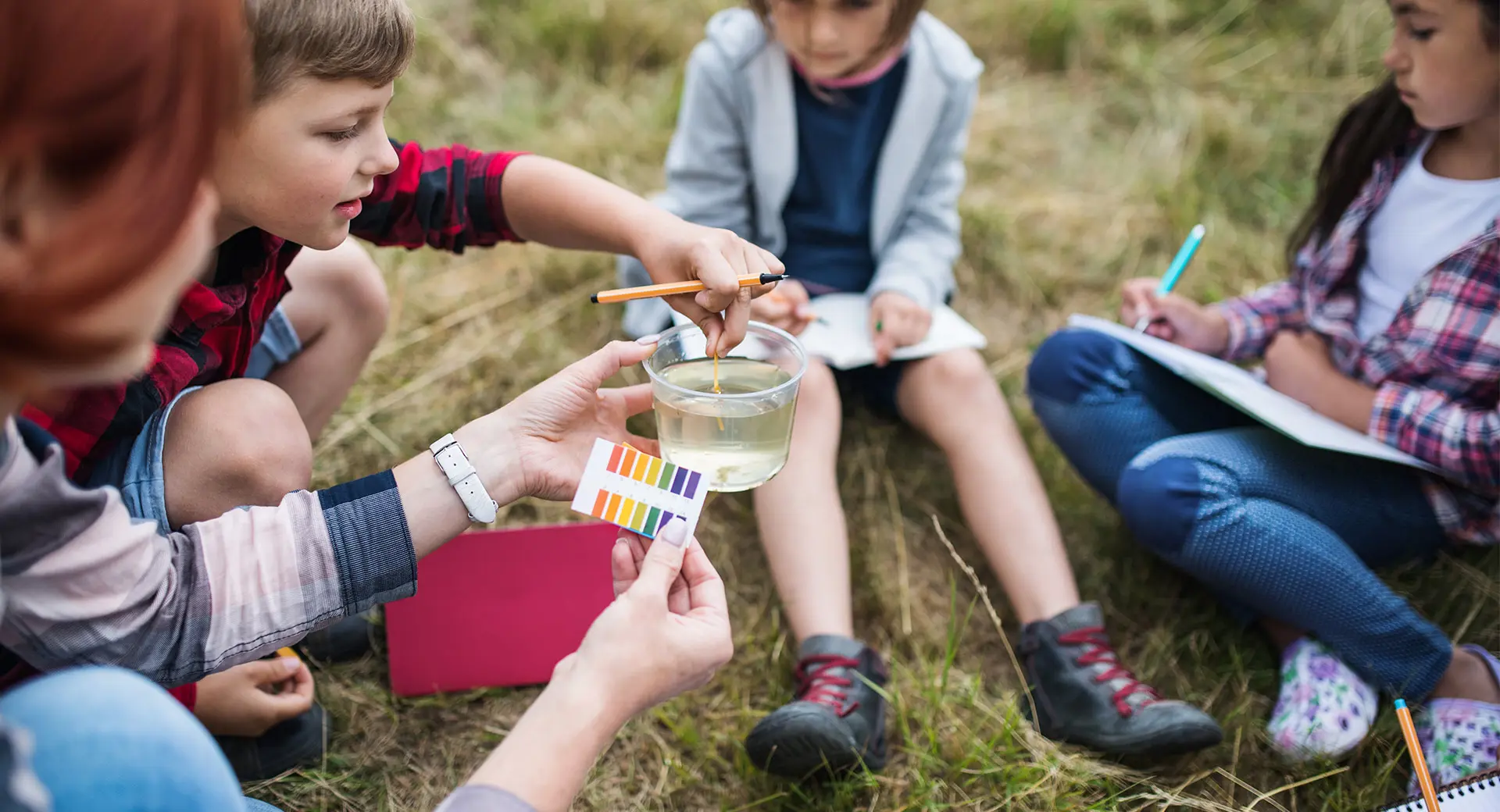 An adult and three children sit in a circle outdoors and test the pH levels of a cup of water