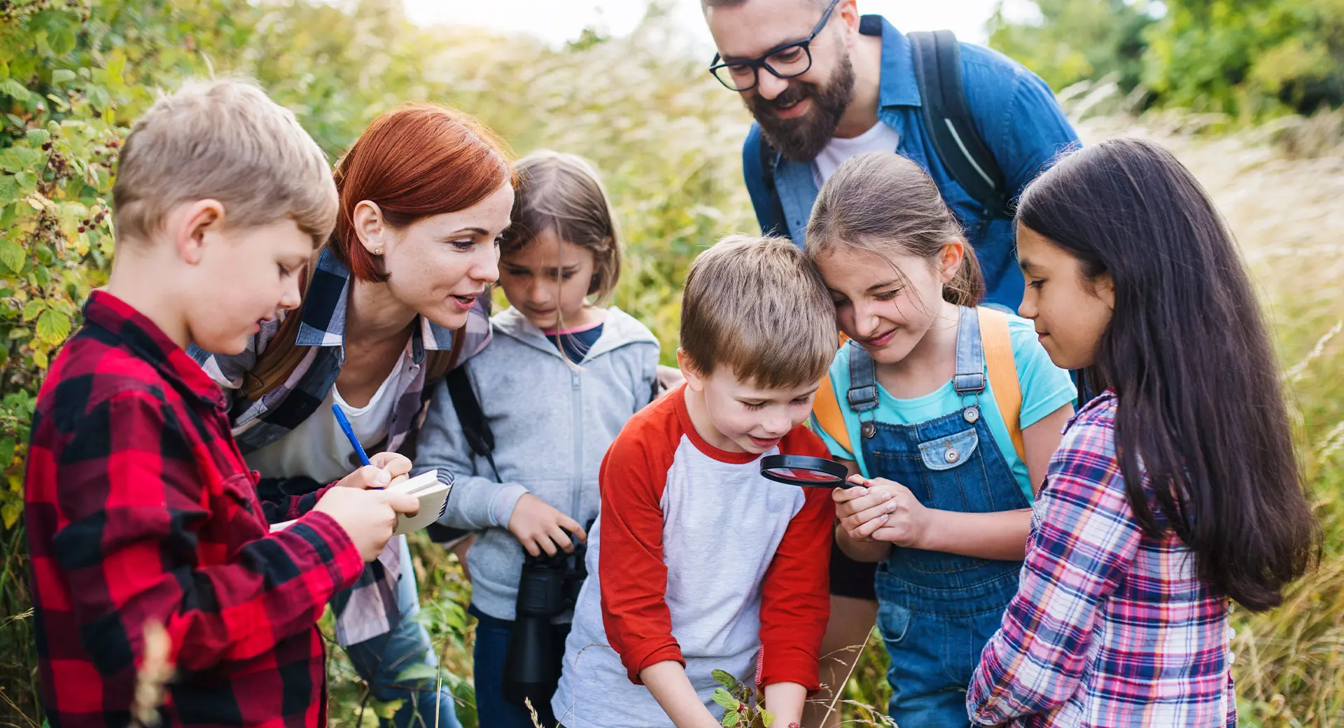 Two adults and five children inspect wildlife with a magnifying glass and take notes in a field